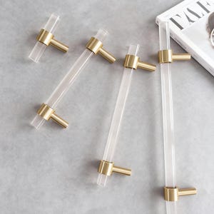 Op de afbeelding: Transparante acryl en messing kastgrepen. De grepen hebben een rechthoekige transparante acryl staaf met messingkleurige uiteinden. De grepen zijn gerangschikt op een lichtgrijze ondergrond. Een tijdschrift en een wijnglas staan op de achtergrond.