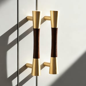 Może przedstawiać: Dwa uchwyty do szafek w stylu mid-century modern. Uchwyty łączą złoty metal i ciemnobrązowe drewno. Uchwyty są zamontowane na białej powierzchni, z rzucanymi cieniami.
