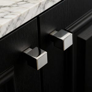 May include: Close-up of a black cabinet with two square, silver cabinet pulls. The cabinet has a marble countertop. The pulls are a modern design, adding a sleek touch to the dark wood.
