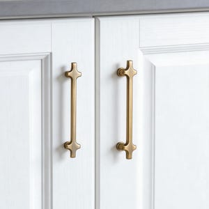 May include: Two gold-colored cabinet pulls with a cross-like design on a white cabinet door. The pulls are vertically oriented and have a long, slender body. The cabinet doors are white with a gray countertop in the background.