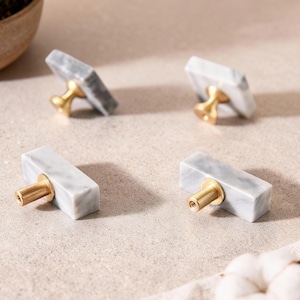 Può includere: Quattro pomelli per mobili con facce rettangolari in marmo grigio e bianco e steli color ottone. I pomelli sono disposti su una superficie chiara. Articolo di ferramenta decorativo.