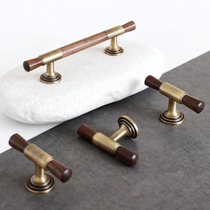 Könnte beinhalten: Eine Sammlung von Schrankbeschlägen mit einer Kombination aus dunklem Holz und antiken Messingoberflächen. Die Griffe und Knöpfe haben ein zylindrisches Design. Die Hardware wird auf einem weißen Stein und einer dunkelgrauen Oberfläche präsentiert.