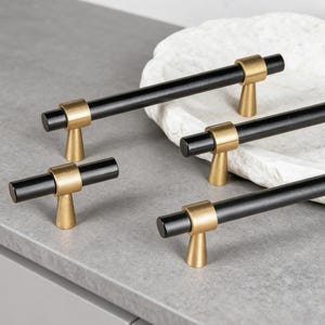 Può includere: Quattro maniglie per mobili nere e dorate disposte su una superficie grigia. Le maniglie hanno un'impugnatura nera cilindrica con dettagli dorati e una base dorata. Adatte per armadi e cassetti.