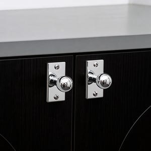 May include: Close-up of two cabinet doors with polished chrome knobs. The knobs are round and mounted on rectangular plates. The cabinet doors are black with a wood grain texture. The top of the cabinet is a dark gray color.