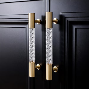 May include: Two cabinet handles with a clear, textured, cylindrical centre and gold-coloured metal ends. The handles are mounted on a black cabinet door. The design features a spiral pattern within the clear cylinder.