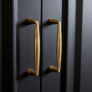 Puede incluir: Primer plano de dos tiradores dorados contra una puerta de armario negra. Los tiradores son largos, curvos y tienen un diseño elegante y moderno. Los tiradores son de metal y tienen un acabado mate.