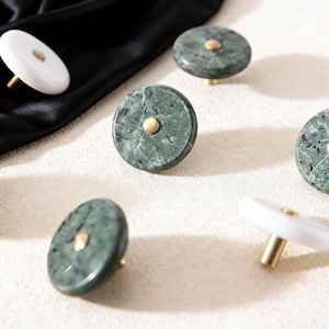 May include: A collection of decorative cabinet knobs. The knobs feature a circular design with a brass-colored center. Some knobs are green marble, while others are white. The knobs are arranged on a light-colored surface.