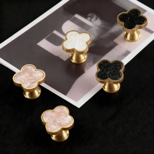 May include: Five clover-shaped cabinet knobs with brass bases. The knobs have white, pink, and black glitter finishes. The knobs are displayed on a black surface and a black and white printed paper. The knobs are for furniture.