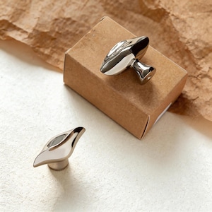May include: Two polished silver leaf-shaped cabinet knobs. One knob is on a small brown cardboard box, and the other is on a white surface. The knobs have a modern, minimalist design, suitable for furniture or decor.