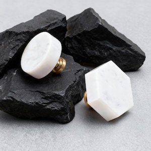 Può includere: Due pomelli per mobili in marmo bianco con finiture in ottone. Un pomello è ovale, l'altro ha una forma esagonale. I pomelli sono esposti su una superficie di roccia nera su sfondo grigio. Arredamento per la casa.