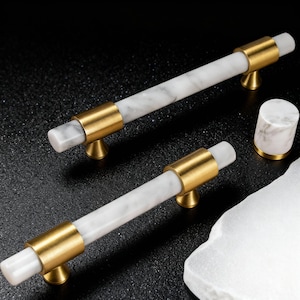 Puede incluir: Dos tiradores de armario con centros blancos efecto mármol y detalles metálicos dorados. Los tiradores son cilíndricos con una banda dorada en el centro y en cada extremo. También se ve un pequeño pomo a juego, junto con un trozo de piedra blanca.