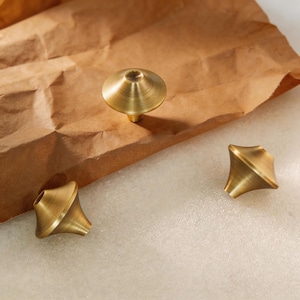 May include: Three brass cabinet knobs in different shapes. One knob is a mushroom shape, and the other two are geometric shapes. The knobs are a warm gold color and are set against a brown paper background and a white surface.