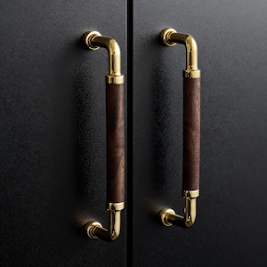 May include: Two cabinet pulls with a gold finish and dark wood center sections are mounted on a black cabinet door. The pulls have a curved design and are evenly spaced, creating a symmetrical appearance.
