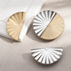 May include: A collection of decorative cabinet hardware. The image shows three pieces: a gold-toned half-circle pull, a silver and gold-toned circular pull, and a silver half-circle pull. All pieces have a fan-like, ridged design.