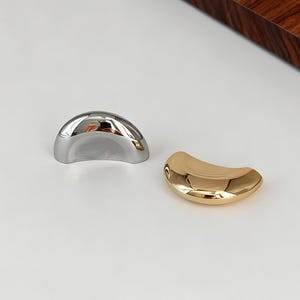 Op de afbeelding: Twee decoratieve meubelgrepen in halve maanvorm, één zilverkleurig en één goudkleurig, met een glad, reflecterend oppervlak. Ze staan op een witte achtergrond met een donkerhouten paneel op de achtergrond.