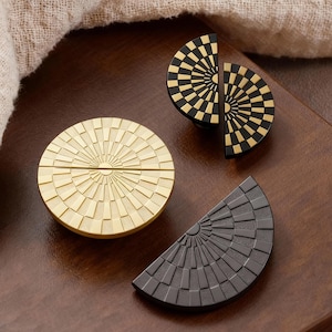 Pode incluir: Uma coleção de ferragens decorativas para armários. Inclui um puxador circular dourado com um design de raios de sol, um puxador semicircular preto e dourado e um puxador semicircular cinza escuro com um padrão geométrico.