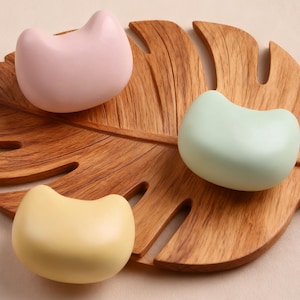 Op de afbeelding: Drie pastelkleurige katvormige objecten in roze, mintgroen en geel zijn gerangschikt op een houten blad-vormige dienblad. De objecten hebben een glad, afgerond ontwerp en de dienblad heeft een natuurlijke houtnerf.