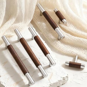 Può includere: Un set di ferramenta per mobili che combina legno e metallo. Le maniglie hanno un centro in legno marrone scuro con estremità in metallo argentato. L'hardware è esposto su una superficie bianca con uno sfondo morbido e strutturato.