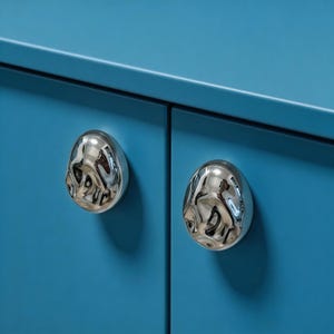 May include: Close-up of two shiny, silver cabinet knobs with a textured, organic design. The knobs are mounted on a bright blue cabinet door, creating a modern and stylish look. Hardware for home decor.
