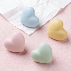 May include: Four pastel-colored heart-shaped objects. One is light blue, another is pink, a third is yellow, and the last is mint green. They are arranged on a pink surface with a piece of white paper. The hearts have a smooth, matte finish.