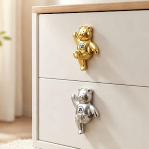 May include: Two drawer knobs shaped like bears, one in a metallic gold finish and one in a metallic silver finish. Each bear is depicted in a waving pose with one arm raised and features a circular emblem on its chest containing the letter W.