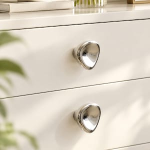 May include: Two polished silver-toned cabinet knobs with an irregular, rounded triangular shape. Each knob features a smooth, reflective metallic surface and is mounted onto a flat, off-white drawer front, providing a clean and functional hardware detail.