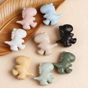 Peut inclure: Une collection de petites figurines en forme de dinosaures, de différentes couleurs pastel : rose, bleu, blanc, beige, jaune, vert et noir. Les figurines sont disposées sur un plateau en bois en forme de feuille.