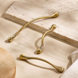 May include: Three curved, matte gold cabinet pulls with rounded, teardrop-shaped ends. Each handle features the black text "Chris&Nicole" printed clearly on one side. The hardware has a smooth, metallic finish and a slender, ergonomic design.