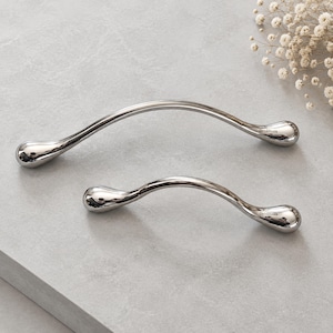 May include: Two polished silver-toned metal cabinet pulls with a curved, elongated shape and rounded ends. Each handle features the engraved text Chris&Nicole on one side. The hardware has a smooth, reflective finish and a sleek, arched design.
