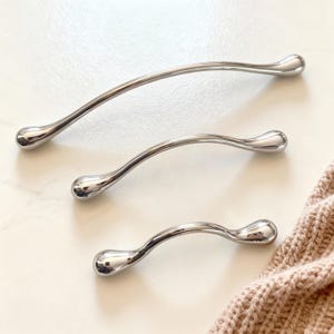 Op de afbeelding: Drie gepolijste, gebogen metalen kastgrepen in verschillende maten. Elke greep heeft een glad, druppelvormig uiteinde. De grepen zijn gerangschikt op een lichtgekleurd oppervlak, met een deel van een beige gebreid item op de achtergrond.