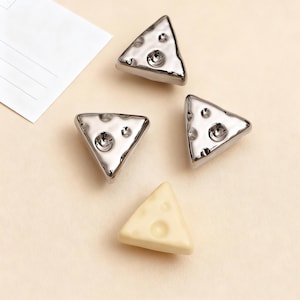 Op de afbeelding: Vier decoratieve objecten in de vorm van kaas. Drie zijn zilverkleurig met gaten en een textuuroppervlak. Eén is lichtgeel met gaten. De objecten zijn gerangschikt op een lichtbeige oppervlak.