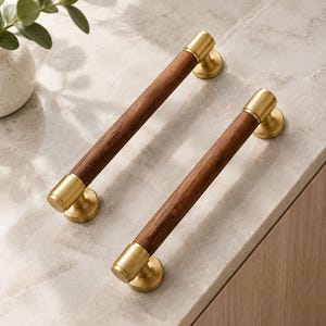 May include: Two cylindrical cabinet pulls featuring dark brown wood handles with gold-toned metal end caps and mounts. The wood surface is engraved with the text Chris & Nicole and the date 05.18.24 in a small, dark, legible font.