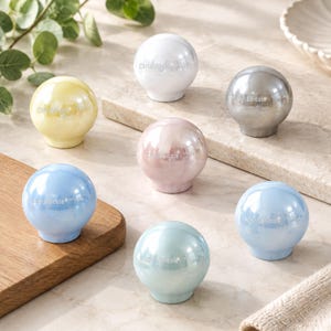 Puede incluir: Siete pomos esféricos para armarios con un acabado brillante e iridiscente en varios colores pastel, incluyendo amarillo, blanco, plata, rosa y tonos de azul. Cada pomo presenta el texto grabado Chris&Nicole en la superficie frontal.
