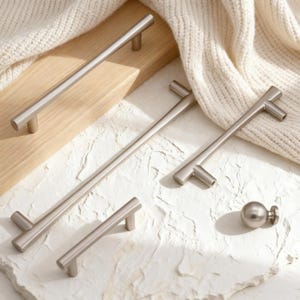 Puede incluir: Un conjunto de herrajes de gabinete de níquel cepillado, que incluyen tiradores de barra y un pomo redondo. El hardware se muestra sobre una superficie de madera clara y un fondo blanco texturizado, con una tela crema. El hardware es para muebles.