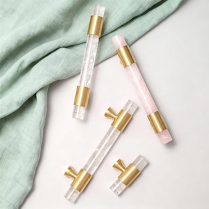 May include: Four decorative cabinet pulls with clear and pink crystal rods and gold-toned metal accents. Two pulls are long, cylindrical handles, and two are shorter with a knob design. The pulls are arranged on a white surface with a green cloth.