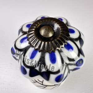 Può includere: Un pomello per mobili in ceramica a forma di fiore smerlato con base bianca, motivi a goccia blu e contorni neri. Il centro presenta una piastra circolare scanalata in metallo scuro. Il testo Chris&Nicole 05.28.24 è visibile.