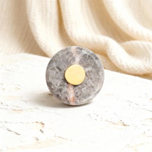 May include: A round, gray marble knob with a gold-colored center. The marble has subtle pink and white veining. This decorative knob is suitable for cabinets, drawers, or furniture.