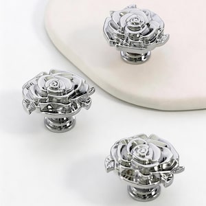 May include: Three polished chrome rose-shaped cabinet knobs. The knobs are highly detailed, with petals and a central bud. They are set against a white background and a curved, off-white surface.