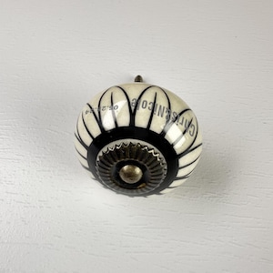 May include: A round ceramic cabinet knob featuring a cream-colored base with black vertical petal-like stripes. The center has a metallic bronze-toned gear-shaped accent. The text Chris & Nicole and the date 05.29.24 are printed on the surface.