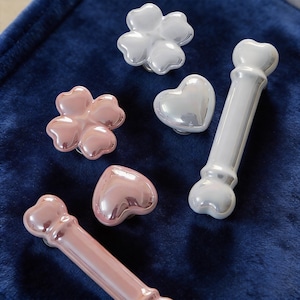 May include: A selection of decorative cabinet knobs in flower, heart, and bone shapes. The knobs are pearlescent in pink and white, set against a dark blue backdrop. These decorative knobs are ideal for adding a touch of elegance.