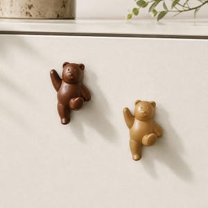 May include: Two bear-shaped cabinet knobs mounted on a flat surface. One knob is dark brown and the other is light tan. Each bear is depicted in a standing pose with one arm raised and one leg bent, featuring smooth, rounded surfaces.