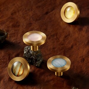 Könnte beinhalten: Dekorative Schrankknöpfe mit Messing-Finish und farbigen, marmorierten Glaseinsätzen. Die Knöpfe sind auf dunkelgrauen Steinsockeln montiert. Die Knöpfe haben ein kreisförmiges Design mit geriffelter Außenkante.