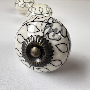 Puede incluir: Un pomo de cerámica redondo para muebles con base blanca y un patrón floral y de hojas negro dibujado a mano. El centro tiene un rosetón de metal color bronce oscuro con un tornillo. La letra M está impresa en un círculo gris en la superficie.
