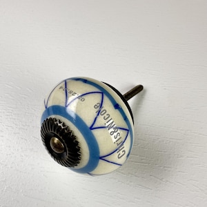 Peut inclure: Un bouton de meuble rond en céramique avec une base crème, orné de motifs de pétales géométriques peints en bleu et du texte Chris&Nicole 08.28.24 imprimé en noir. Le bouton possède un centre métallique sombre dentelé et une vis.