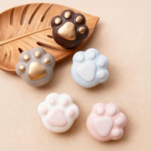 Op de afbeelding: Vijf pootvormige decoratieve items in verschillende kleuren: bruin met gouden accenten, grijs met gouden accenten, lichtblauw, wit met roze accenten en roze. De items zijn gerangschikt op een houten blad-vormige dienblad.