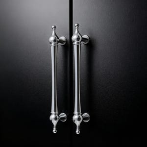 Puede incluir: Dos tiradores de armario cromados pulidos con un diseño decorativo y alargado. Los tiradores están montados sobre una superficie oscura y texturizada, creando un contraste entre el metal brillante y el fondo mate. Los tiradores tienen un estilo clásico y elegante.