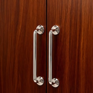 May include: Two polished chrome cabinet door pulls with a curved handle design. The pulls are mounted on a dark brown wooden cabinet door. The handles are vertically oriented and have a circular base.