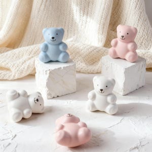 Puede incluir: Colección de figuras de osos de peluche de cerámica en varios colores: azul claro, rosa y blanco. Los osos están sentados o tumbados sobre pedestales blancos en forma de cubo. Cada oso tiene una cara simple y sonriente.