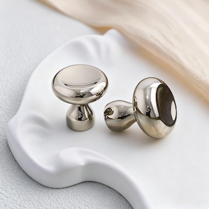 Op de afbeelding: Drie glanzende, zilveren kastknoppen met een paddenstoelachtig ontwerp worden gepresenteerd op een witte, golvende tray. De knoppen hebben een glad, reflecterend oppervlak, waarbij het licht highlights en schaduwen creëert. Deze knoppen zijn waarschijnlijk voor meubels of decoratieve doeleinden.