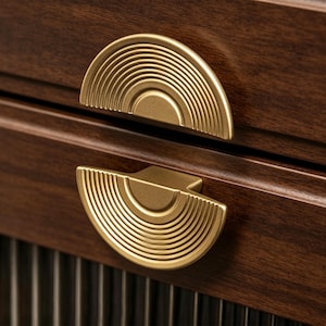 May include: Two semi-circular drawer pulls made of brushed gold metal, each featuring a series of concentric circular grooves. The pulls are mounted on a dark brown wooden cabinet surface with a visible grain and vertical slat detailing below.